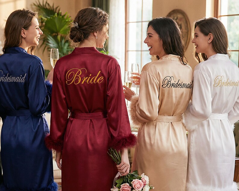 May include: Four women in satin robes of navy, burgundy, champagne, and white. The burgundy robe has "Bride" embroidered in gold, the others say "Bridesmaid". The robes have feather trim. The women are holding glasses of champagne.