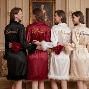 May include: Four women wearing satin robes in black, burgundy, white, and champagne. Each robe has feather trim on the cuffs and hem. The robes are embroidered with the words "Bride" and "Bridesmaid" in gold or black thread.
