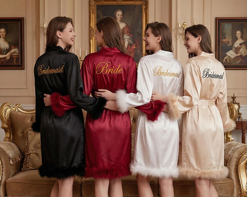 May include: Four women wearing satin robes in black, burgundy, white, and champagne. Each robe has feather trim on the cuffs and hem. The robes are embroidered with the words "Bride" and "Bridesmaid" in gold or black thread.