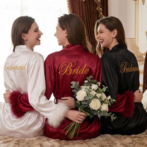 May include: Three women in satin robes: white with "Bridesmaid" in gold, burgundy with "Bride", and black with "Bridesmaid". Each robe has feather trim. A bouquet of white flowers is held.