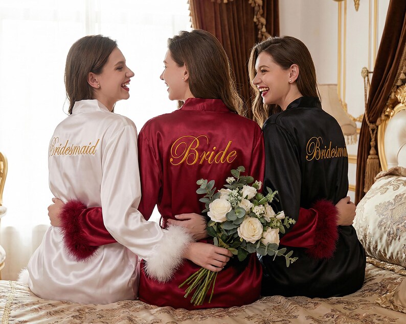 May include: Three women in satin robes: white with "Bridesmaid" in gold, burgundy with "Bride", and black with "Bridesmaid". Each robe has feather trim. A bouquet of white flowers is held.
