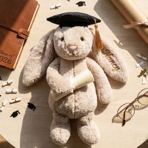 May include: A soft toy bunny wearing a graduation cap and holding a scroll. The bunny's ear is embroidered with the name "Nancy". A brown leather journal with "Class of 2026" is visible. Graduation-themed confetti and eyeglasses are scattered around.