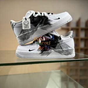 Puede incluir: Zapatillas blancas con diseño de pinceladas negras y grises. Presentan un logotipo de Nike negro y una etiqueta que dice "Happy Smile". Los zapatos tienen cordones blancos y una suela blanca.