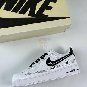 Könnte beinhalten: Weißer Nike Air Force 1 Sneaker mit schwarzen Akzenten, einschließlich des Nike-Swoosh, der Schnürsenkel und der Ferse. Der Schuh verfügt über individuelle Grafiken und Text, einschließlich des Wortes "AIR" auf der Zwischensohle. Der Schuh wird mit der Originalverpackung präsentiert.