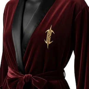 May include: A burgundy velvet robe with black satin lapels and a matching tie belt. The robe features a gold embroidered emblem on the chest. The robe is displayed on a mannequin.