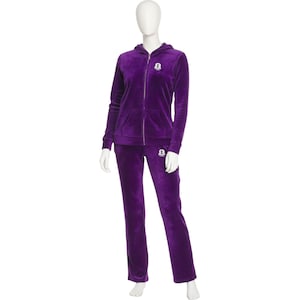 May include: A two-piece purple velour tracksuit displayed on a mannequin. The set includes a zip-up hoodie with a hood and front pockets, and matching straight-leg pants. A small emblem is visible on the chest and hip.