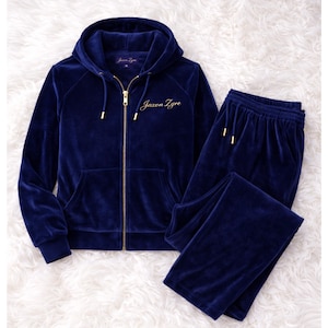 May include: A navy blue velour tracksuit set. The set includes a zip-up hoodie with a hood and front pockets, and matching tracksuit bottoms. The brand name "Jaxon Zyre" is embroidered in gold on the hoodie. The tracksuit is displayed on a white, fluffy surface.