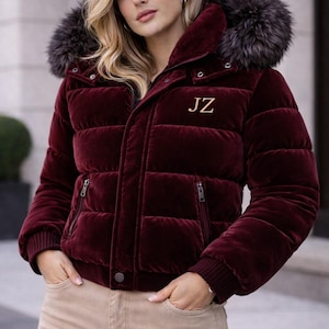 Op de afbeelding: Een bordeauxrode fluwelen pufferjas met een met bont afgezette capuchon. De jas heeft een gewatteerd design, een ritssluiting en de initialen "JZ" in goud geborduurd. Het model draagt een beige broek.