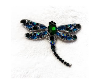 May include: A silver dragonfly brooch featuring a central green gem and wings embellished with blue and black crystals. The dragonfly's body is detailed with black crystals, creating a sparkling accessory.