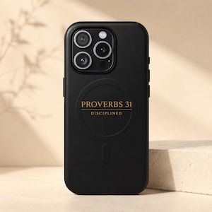 May include: A matte black smartphone case featuring a triple-lens camera cutout at the top. Centered on the back is a circular magnetic charging ring with the gold text PROVERBS 31 and the word DISCIPLINED printed directly underneath the text.