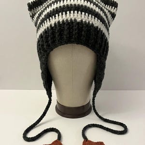 May include: A handmade crochet hat with cat ears, featuring alternating stripes of black and white. The hat has long earflaps with star-shaped embellishments in a rust color. The hat is displayed on a mannequin head.