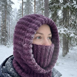 Peut inclure: Une cagoule violette tricotée avec une texture côtelée, couvrant la tête et le cou. La cagoule est portée à l'extérieur dans un cadre forestier enneigé, offrant chaleur et protection contre le froid.