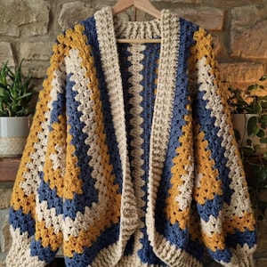 Peut inclure: Cardigan fait main, ouvert sur le devant, en motif granny square. La veste en crochet présente un motif color block avec des carrés crème, bleu marine et jaune moutarde. Le cardigan est présenté sur un cintre en bois contre un mur en pierre.