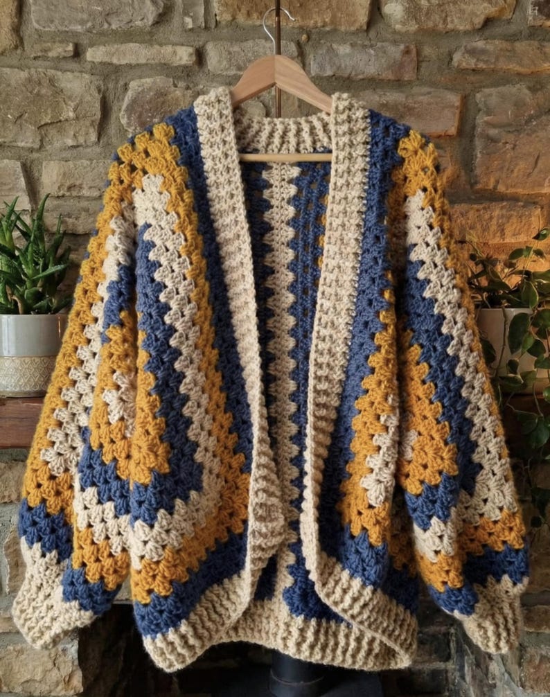 Peut inclure: Cardigan fait main, ouvert sur le devant, en motif granny square. La veste en crochet pr&eacute;sente un motif color block avec des carr&eacute;s cr&egrave;me, bleu marine et jaune moutarde. Le cardigan est pr&eacute;sent&eacute; sur un cintre en bois contre un mur en pierre.