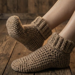 Può includere: Un paio di pantofole all'uncinetto marroni fatte a mano. Le pantofole hanno un polsino a coste e un design a trama aperta strutturata. L'immagine mostra le pantofole su una superficie di legno.