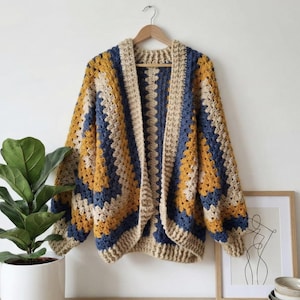 Peut inclure: Cardigan en crochet fait main, motif granny square. Le pull sans fermeture pr&eacute;sente un design color block avec des nuances de beige, bleu marine et jaune moutarde. Le v&ecirc;tement est pr&eacute;sent&eacute; sur un cintre en bois contre un mur blanc.