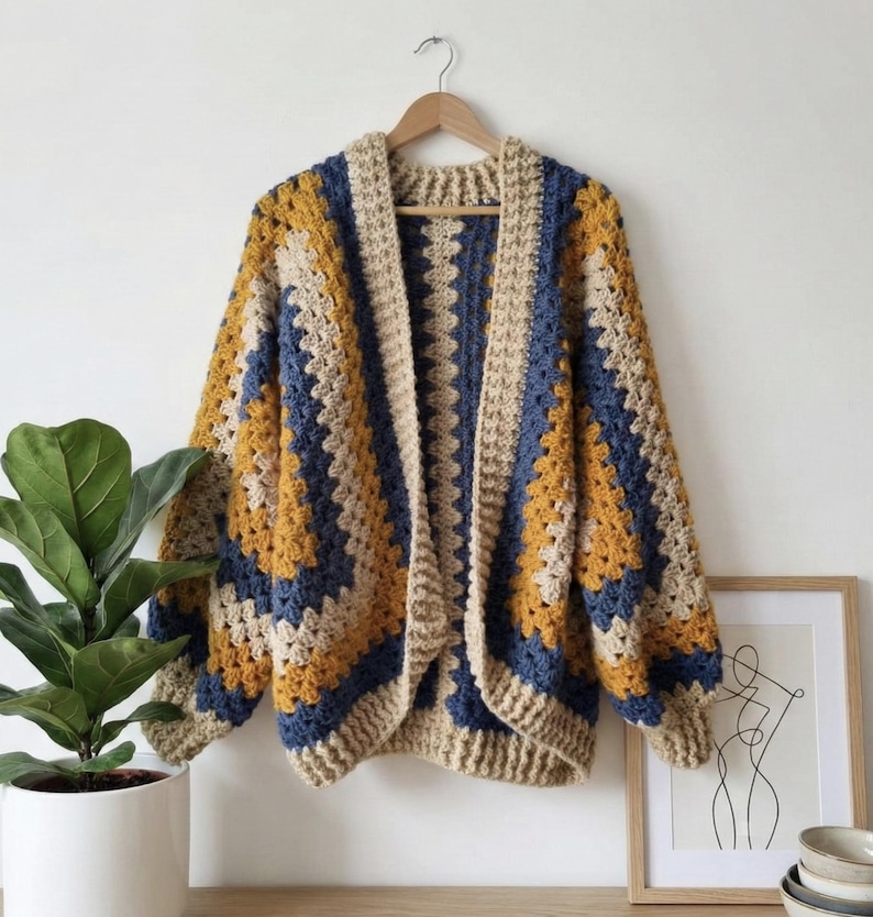 Peut inclure: Cardigan en crochet fait main, motif granny square. Le pull sans fermeture pr&eacute;sente un design color block avec des nuances de beige, bleu marine et jaune moutarde. Le v&ecirc;tement est pr&eacute;sent&eacute; sur un cintre en bois contre un mur blanc.