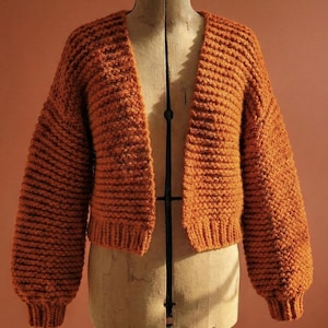 Puede incluir: Un cárdigan naranja con frente abierto, con dobladillo y puños acanalados. El suéter se exhibe en un maniquí vintage. Las mangas son largas y el tejido tiene una apariencia texturizada.