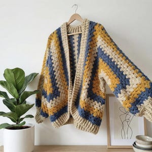 Peut inclure: Un cardigan en crochet fait &agrave; la main, &agrave; devant ouvert, avec un motif g&eacute;om&eacute;trique. Le sch&eacute;ma de couleurs comprend des rayures beiges, jaune moutarde et bleu marine. Le cardigan est suspendu &agrave; un cintre en bois contre un mur blanc.