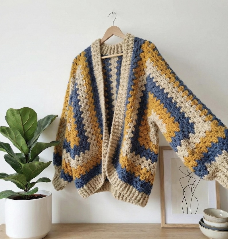 Peut inclure: Un cardigan en crochet fait &agrave; la main, &agrave; devant ouvert, avec un motif g&eacute;om&eacute;trique. Le sch&eacute;ma de couleurs comprend des rayures beiges, jaune moutarde et bleu marine. Le cardigan est suspendu &agrave; un cintre en bois contre un mur blanc.