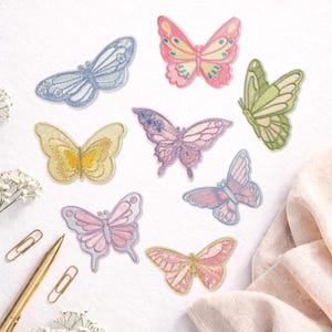 May include: A collection of embroidered butterfly patches in various colors, including pink, blue, yellow, and green. The patches feature detailed stitching and are arranged on a white surface. Some patches have delicate floral accents.