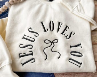 May include: Cream-colored sweatshirt with the embroidered phrase "JESUS LOVES YOU" in black, arched across the front. A black bow design is centered below the text. The sweatshirt is folded, with a portion of the sleeve visible.