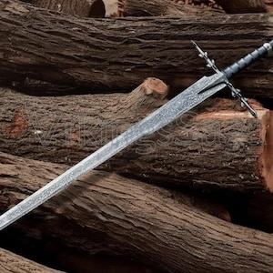 May include: A detailed image of a long, silver sword with a black handle and crossguard. The sword is resting on a pile of rough-hewn logs, showcasing its intricate design and craftsmanship. The blade has a textured surface.