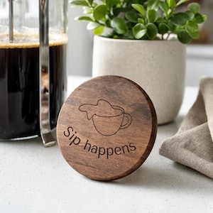 Peut inclure: Un couvercle circulaire en bois foncé présentant un dessin au trait gravé d'une tasse de café avec une éclaboussure et le texte "Sip happens" gravé sous le motif. Le couvercle en bois a une finition lisse et un grain naturel visible.