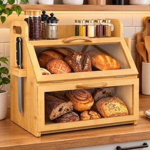 May include: A two-tier bamboo bread box with clear acrylic windows and handles. The top shelf holds glass spice jars, while the interior contains various loaves of bread. A knife is stored in a side slot on the left exterior panel.