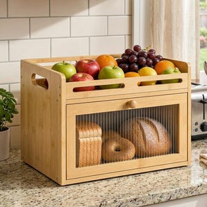 May include: A rectangular bamboo bread box with a drop-down door featuring a ribbed glass panel. The top surface serves as a storage tray holding a variety of fresh fruit, including red and green apples, oranges, and a cluster of purple grapes.