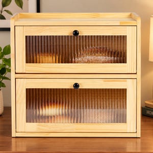 May include: A two-tier wooden bread box featuring light-toned wood construction and two drop-down doors. Each door is equipped with a small black circular knob and a clear, vertically ribbed glass panel that partially obscures the interior contents.