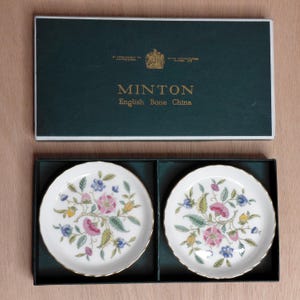 May include: Two small, white English bone china plates with floral designs in a dark green box. The plates feature pink, blue, and yellow flowers with green leaves. The box is dark green with the word "MINTON" and "English Bone China" printed on it.