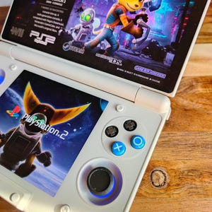 Può includere: Una console di gioco portatile Nintendo 3DS bianca che mostra uno schermo di gioco con un personaggio dei cartoni animati. La console ha pulsanti blu e neri e un D-pad. Lo schermo superiore mostra il titolo e i personaggi del gioco. La console è su una superficie di legno.