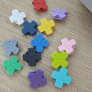 Könnte beinhalten: Eine Sammlung von dreizehn D-Pad-Controller-Tasten in verschiedenen Farben wie Rot, Weiß, Blau, Gelb, Grau, Schwarz, Grün, Lila, Pink und Hellblau. Jede Taste hat eine Kreuzform mit eingeprägten Richtungspfeilen auf der Oberfläche.