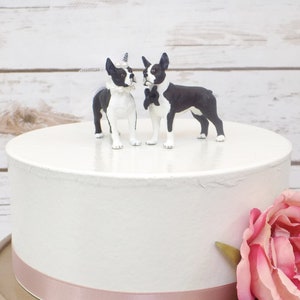May include: A white wedding cake with a pink ribbon around the bottom tier. Two black and white Boston Terrier figurines are on top of the cake, one wearing a veil and the other a bow tie.
