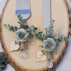 May include: A set of two silver cake knives with wooden handles decorated with greenery, a light blue flower, and a heart-shaped wooden tag. The tag on the left knife says "Jonathan & Grace" and the tag on the right knife says "July 1, 2021".
