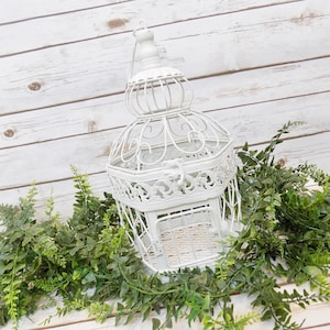 May include: A white decorative birdcage with intricate scrollwork and a handle, set against a white wooden background. Green leafy garland frames the cage, adding a natural touch. The cage is approximately 12 inches tall.