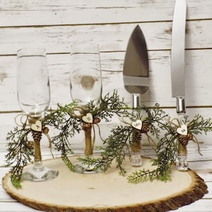 May include: A rustic wedding cake serving set with two champagne flutes, a cake server, and a knife. The set is decorated with greenery, pinecones, and wooden hearts. The flutes and knife handles are wrapped in twine.
