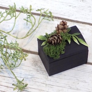 May include: A small black wooden box with a hinged lid, decorated with pine cones, moss, and green leaves. The box is sitting on a white wooden surface.
