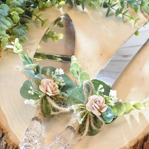 May include: A set of two cake cutting knives with clear handles and silver blades. Each knife is decorated with a bouquet of faux greenery, a pink rose, and twine.
