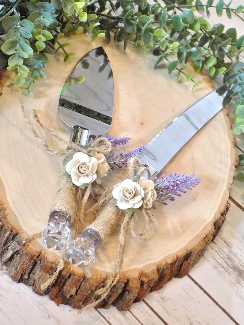 May include: A set of two cake cutting knives with rustic brown twine and white fabric flowers. The knives are on a wooden slice with lavender sprigs.