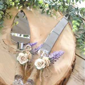 May include: A set of two silver cake cutting knives with rustic brown burlap and white flower accents. The knives are on a wooden slice with green foliage.