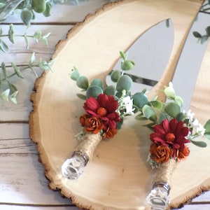 May include: A set of two cake cutting knives with rustic floral accents. The knives have clear glass handles and are decorated with burgundy and orange flowers, greenery, and burlap ribbon.