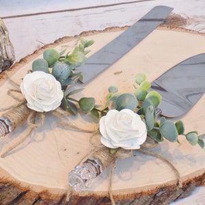 May include: A set of two cake cutting knives with white roses and greenery tied around the handles. The knives are on a wooden slice.