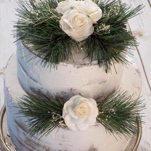May include: A two-tiered white cake decorated with white roses and green pine needles. The cake is on a silver stand, with a rustic, textured frosting. The top tier features three roses, while the bottom tier has one.