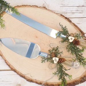 May include: A set of two silver cake knives with wooden handles decorated with pine cones, greenery, and twine. The handles are adorned with small wooden hearts that say "Mr." and "Mrs."
