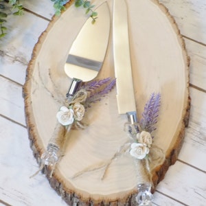 May include: A rustic wooden cake server set with two silver knives decorated with burlap, white flowers, and lavender sprigs.