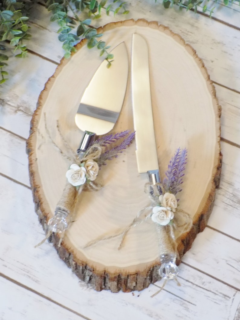 May include: A rustic wooden cake server set with two silver knives decorated with burlap, white flowers, and lavender sprigs.
