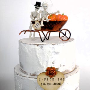 May include: A white wedding cake with a skeleton couple riding in a wheelbarrow with pumpkins. The cake topper says "T&C" and the cake has a wooden heart-shaped sign that says "I PICK YOU 10.25.2025".