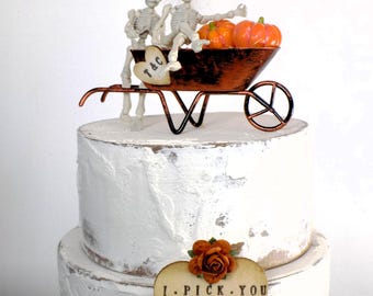 May include: A white wedding cake with a skeleton couple riding in a wheelbarrow with pumpkins. The cake topper says "T&C" and the cake has a wooden heart-shaped sign that says "I PICK YOU 10.25.2025".